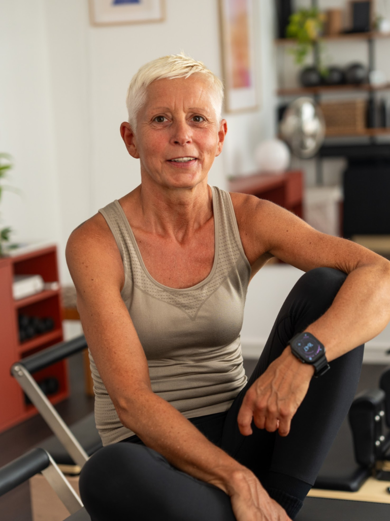 Frau mittleren Alters mit kurzen blonden Haaren sitzt auf dem Reformer und lächelt in die Kamera.